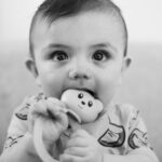 A cute baby bites a toy while sitting in a relaxed indoor setting, creating a charming portrait.